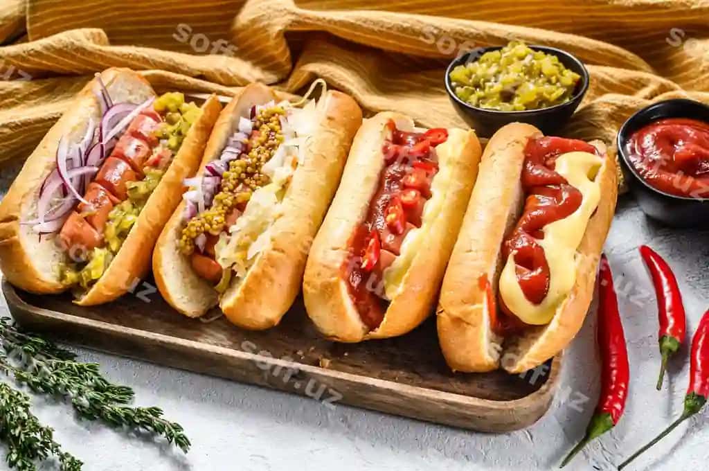 Turli xil toppinqar bilan to'ldirilgan hot doglar diskasi, gurtli va qurdiu go'shtli kolbasali mumtostilik hot doglar, oq fonda yuqori ko'rinish. | Hot dogs fully loaded with assorted toppings tray delicious hot dogs with pork beef sausages white background top view