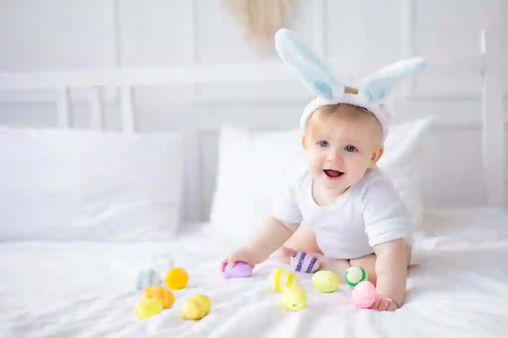 Quyon quloqli shirin bolakay rangli tuxum oq karavotda uyda tuxum yeyayotgan sarg‘ish sochli chaqaloq baxtli Pasxa bayrami tushunchasi | Cute baby boy with bunny ears colorful eggs white bed home playing eating eggs little blonde baby happy easter concept